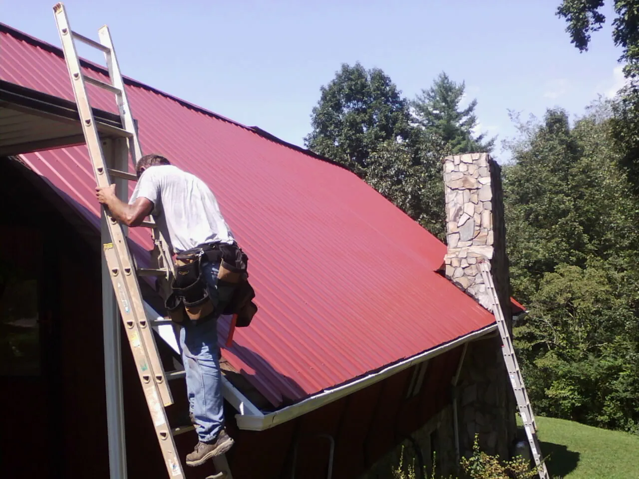 Roofer on a ladder beside a red metal roof and stone chimney — Soffit & Fascia Repair by Roofing Brothers in Crystal Lake